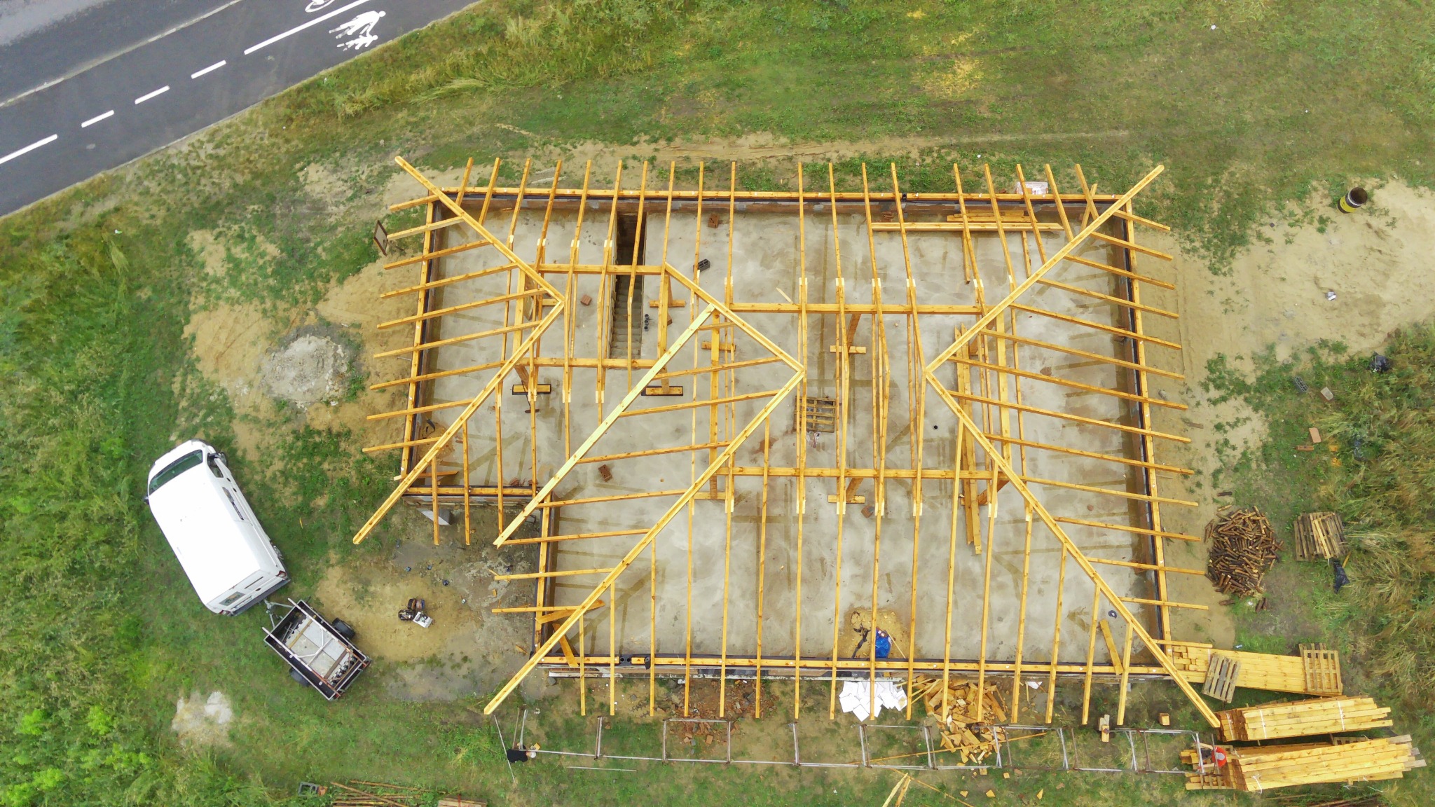 Widok z drona na budowę domu z widoczną konstrukcją drewnianą dachu, biały bus z przyczepką zaparkowany obok, w tle droga asfaltowa.