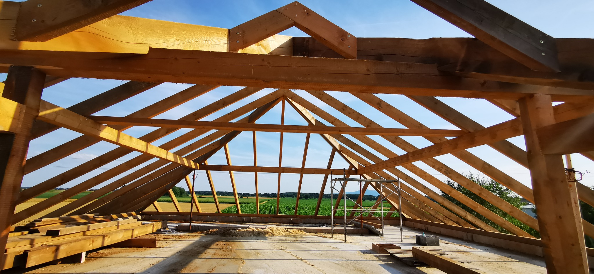 Konstrukcja drewniana dachu w trakcie montażu, widok na otwartą przestrzeń i pola uprawne przez szkielet dachu, ze stertą drewna na pierwszym planie i rusztowaniem w środku kadru.
