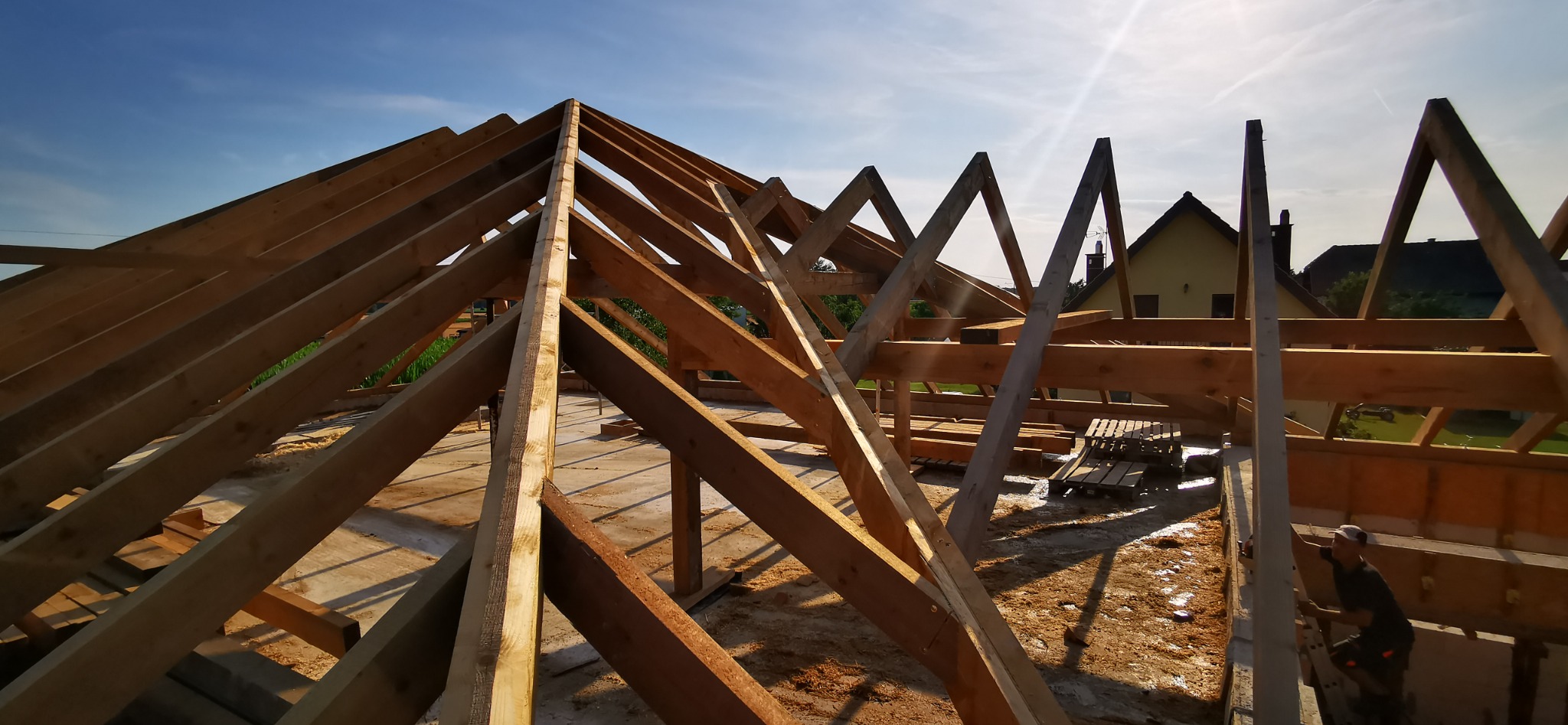 Konstrukcja drewniana dachu w trakcie budowy, widoczne elementy więźby dachowej na tle błękitnego nieba z widocznym budynkiem mieszkalnym w tle.