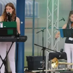 Dwie młode kobiety występują na scenie: jedna gra na keyboardzie Yamaha, druga na skrzypcach. Na pierwszym planie leży trąbka.