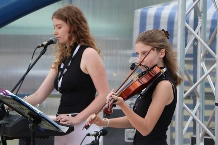 Dwie młode kobiety grające muzykę na żywo: jedna na keyboardzie z mikrofonem, druga na skrzypcach z mikrofonem przypiętym do skrzypiec, na tle metalowej konstrukcji.