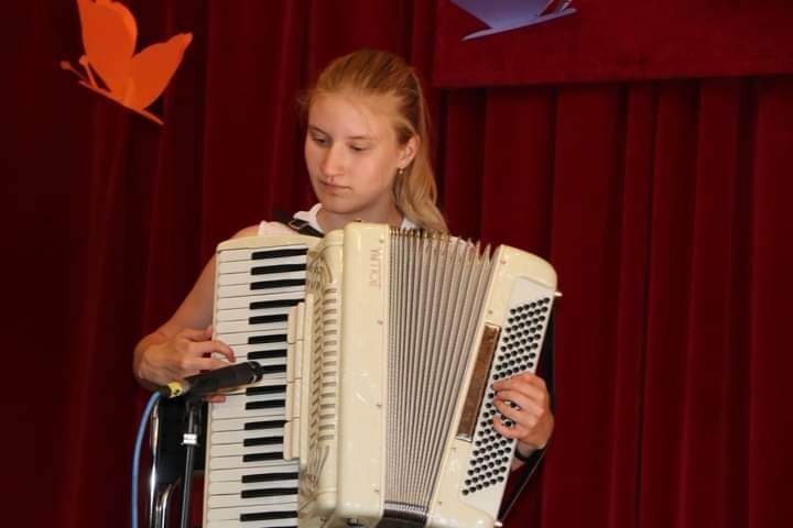 Młoda kobieta grająca na białym akordeonie marki 'Bollma' na tle czerwonej zasłony i dekoracji z motylem.