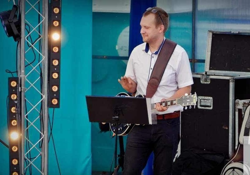 Gitarzysta w białej koszuli, grający na scenie z oświetleniem LED i czarnym case na instrumenty w tle.