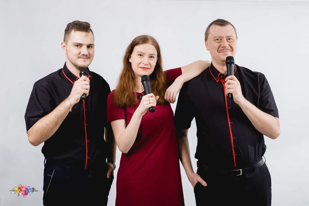 Trzyosobowy zespół muzyczny w formalnych strojach, z mikrofonami, pozujący na białym tle. Kobieta z ręką opartą na ramieniu mężczyzny po prawej.