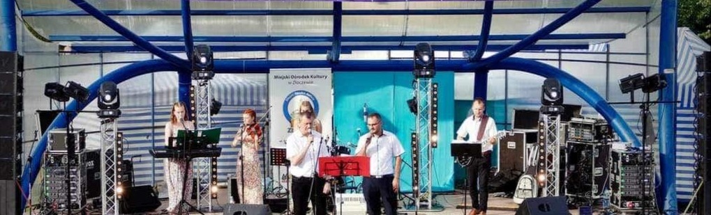 Występ zespołu muzycznego na plenerowej scenie, widoczni muzycy grający na instrumentach (keyboard, skrzypce, gitara), wokaliści oraz nagłośnienie i oświetlenie sceniczne, w tle baner Miejskiego...