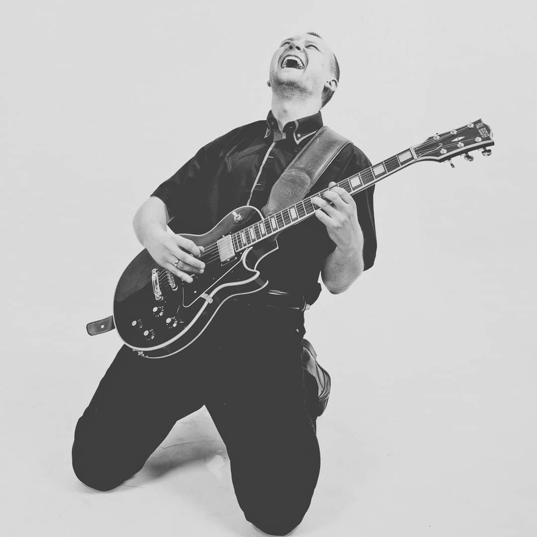 Czarno-białe zdjęcie rozentuzjazmowanego gitarzysty grającego na elektrycznej gitarze, klęczącego na jednym kolanie z głową odchyloną do tyłu i szeroko otwartymi ustami.