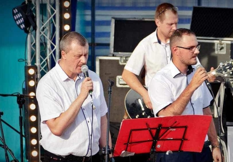 Trzech mężczyzn w białych koszulach występuje na scenie: jeden śpiewa do mikrofonu, drugi gra na gitarze, trzeci stoi w tle. Na scenie widoczne oświetlenie i czerwony pulpit na nuty.