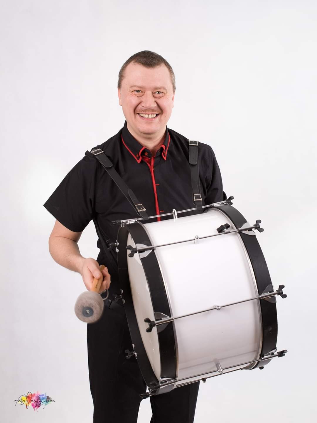 Uśmiechnięty mężczyzna w czarnej koszuli z czerwonym wykończeniem trzyma duży biały bęben z czarnymi obręczami i pałką z futrzaną końcówką, zawieszony na szelkach.