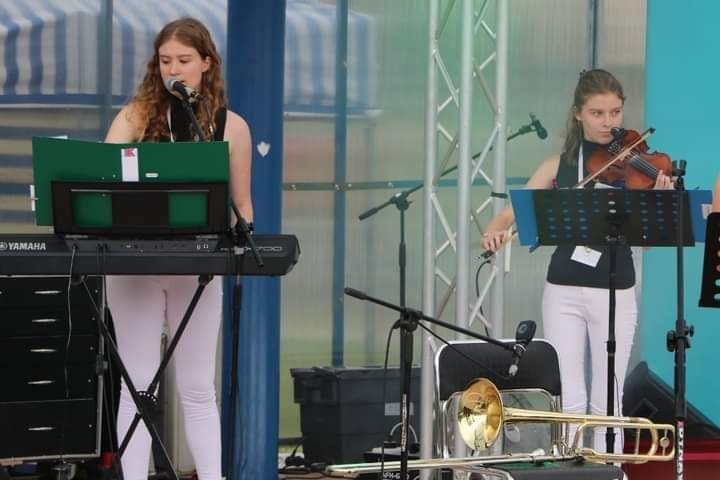 Dwie młode kobiety występują na scenie: jedna gra na keyboardzie Yamaha, druga na skrzypcach. Na pierwszym planie leży trąbka.