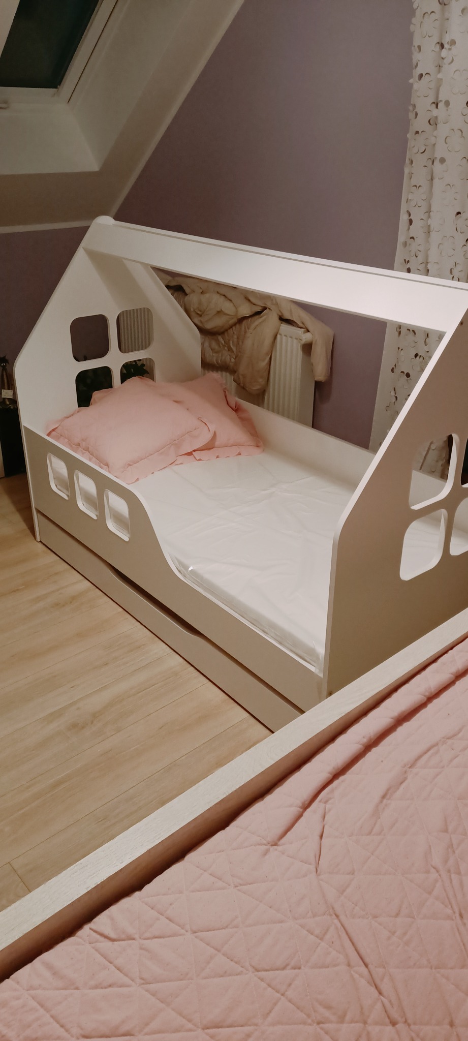 Białe łóżko dziecięce w kształcie domku z różową pościelą i poduszkami, widoczne na tle pokoju z drewnianą podłogą i ścianą w odcieniach fioletu.