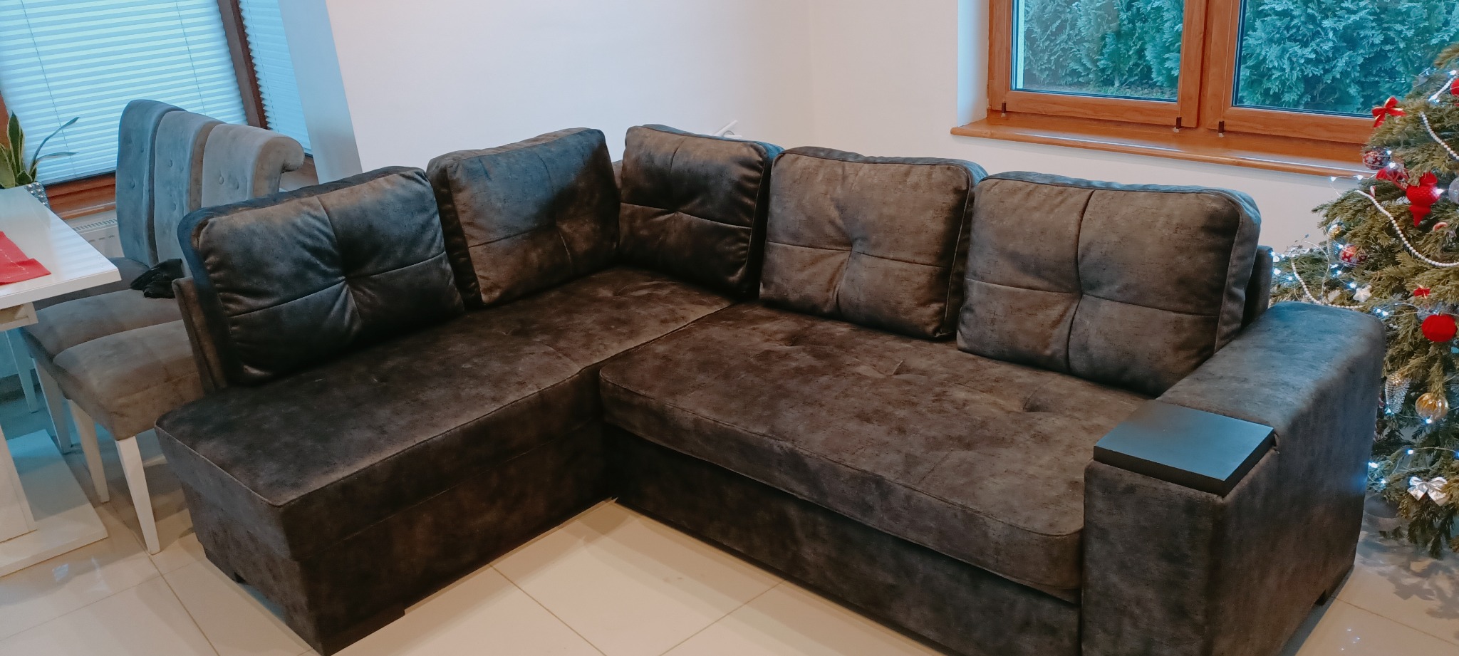 Narożna sofa w odcieniach szarości z kwadratowymi poduszkami, obok okna i choinki świątecznej.