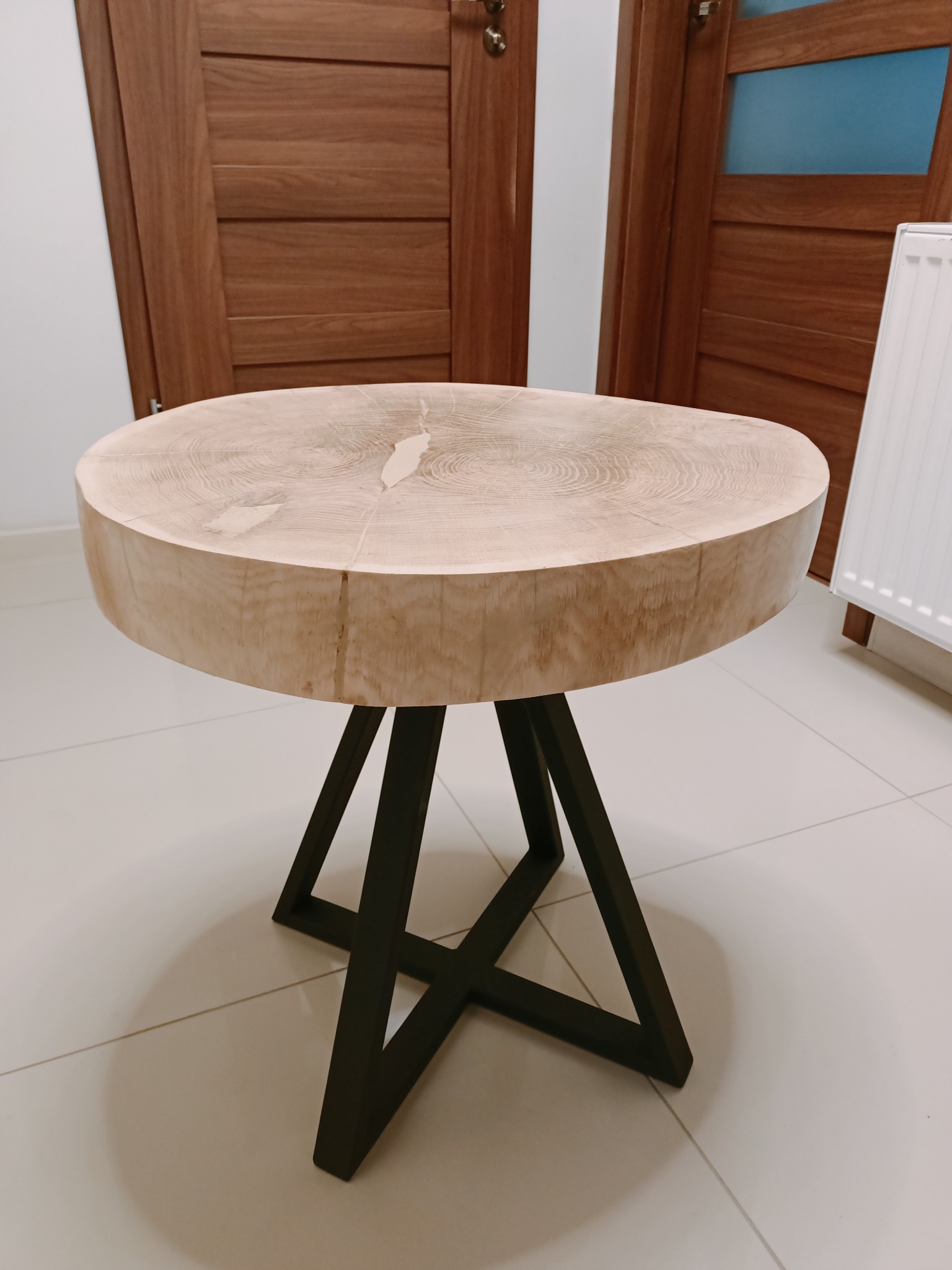 Okrągły stolik kawowy z blatem z jasnego drewna z widocznymi słojami i czarnymi, metalowymi nogami w kształcie litery X, na tle drewnianych drzwi i białej ściany.