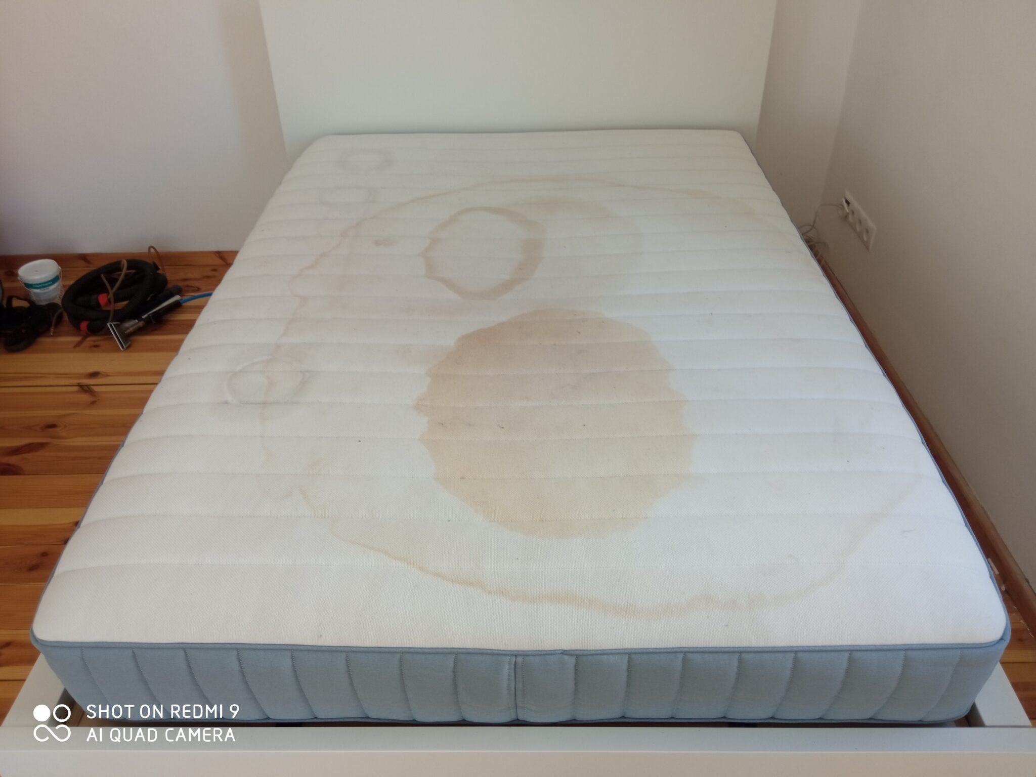 Materac z widocznymi, dużymi plamami przed czyszczeniem, obok leżą narzędzia do czyszczenia tapicerki.