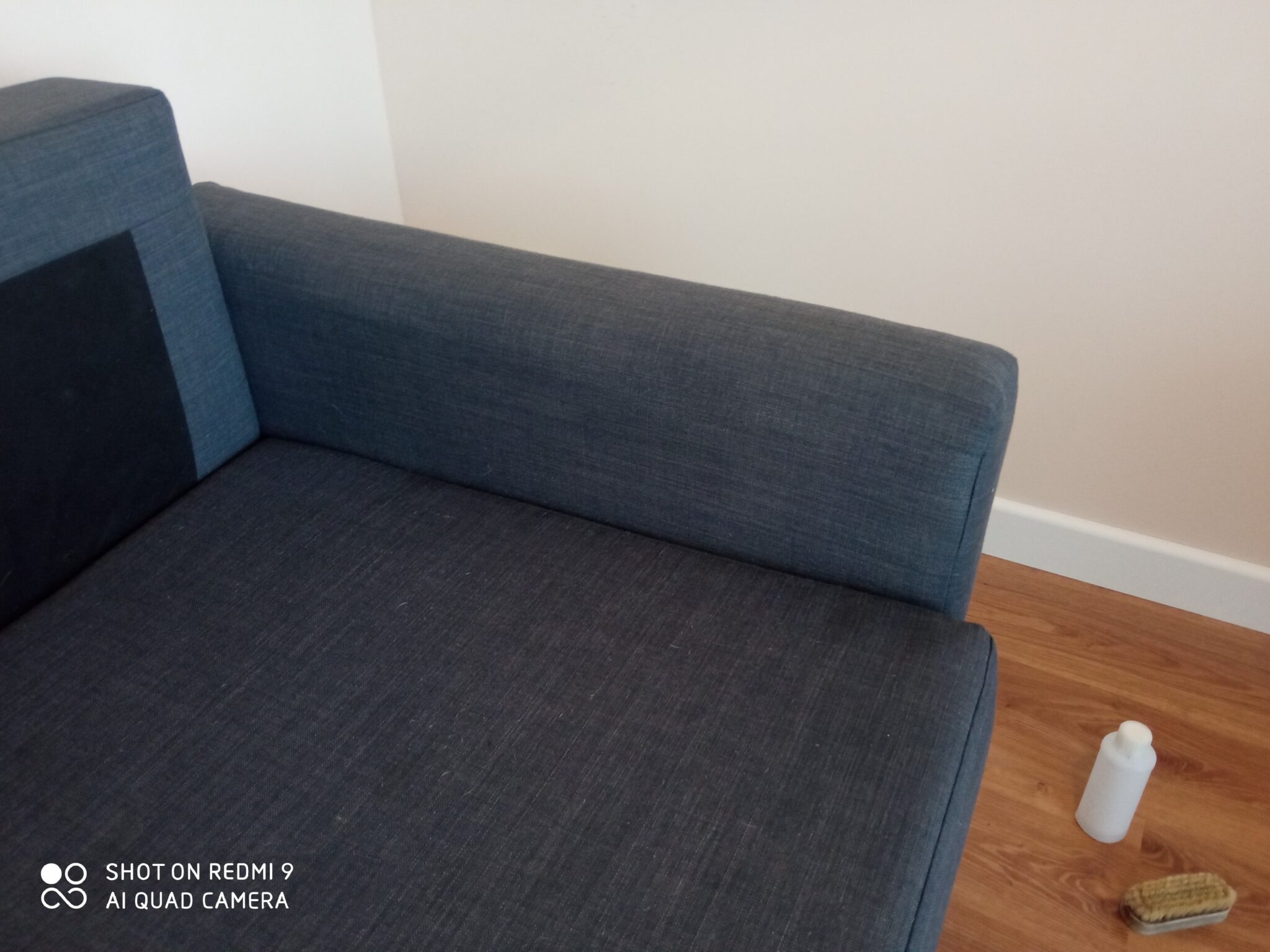 Fragment niebieskiej sofy z ciemnym panelem bocznym, obok butelka środka czyszczącego i szczotka na drewnianej podłodze, na tle białej ściany.