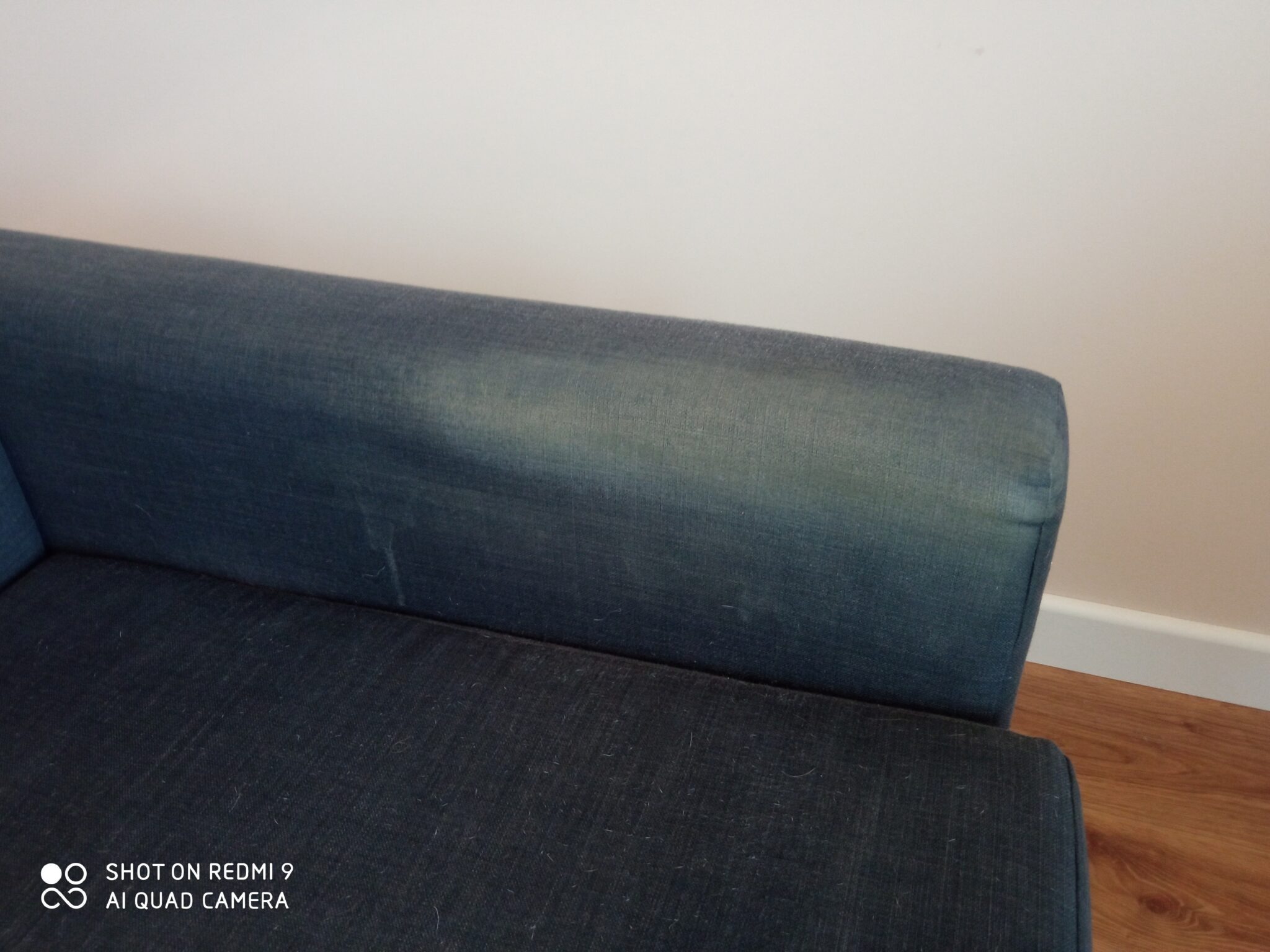 Fragment ciemnoniebieskiej kanapy z widocznymi śladami użytkowania i drobnymi zabrudzeniami na tapicerce, na tle beżowej ściany i drewnopodobnej podłogi.