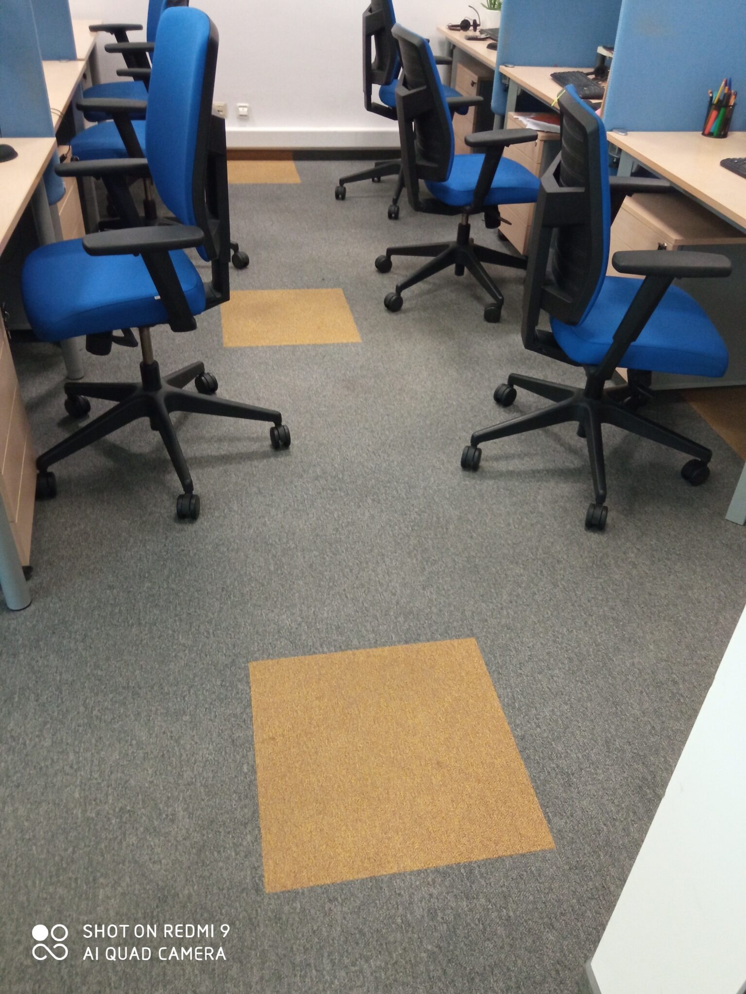Szary dywan w biurze z żółtymi kwadratami, pomiędzy biurkami z niebieskimi krzesłami obrotowymi.
