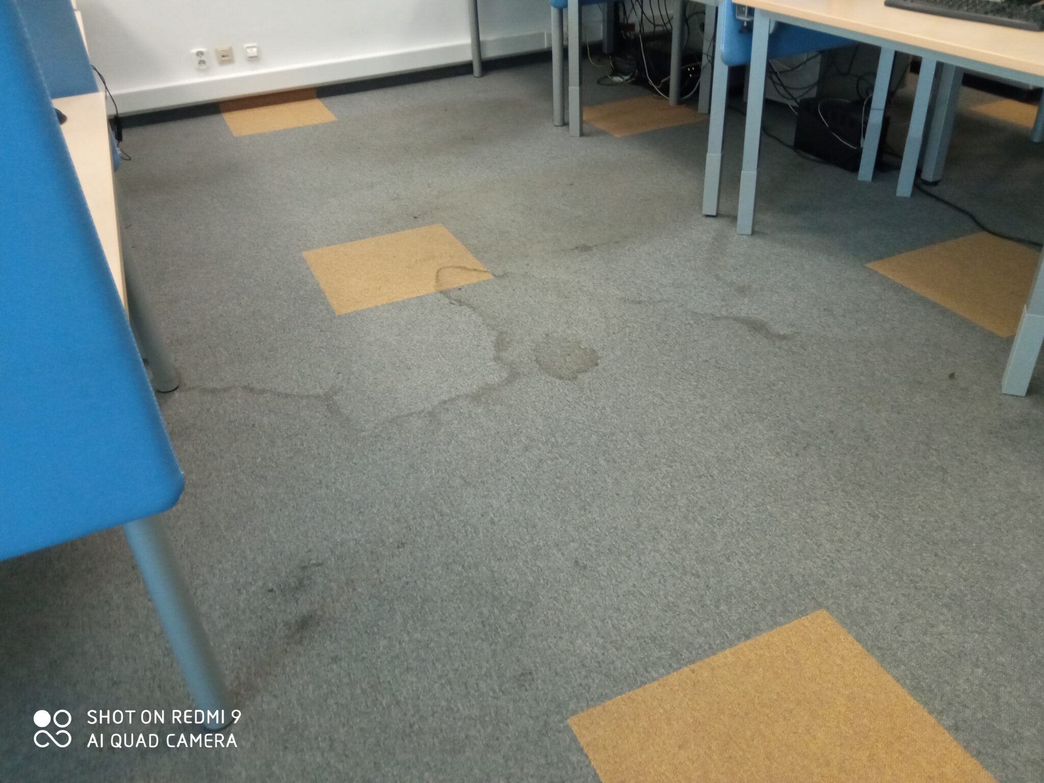 Szary dywan w biurze z plamami i żółtymi kwadratami, widoczne nogi biurek i krzeseł.