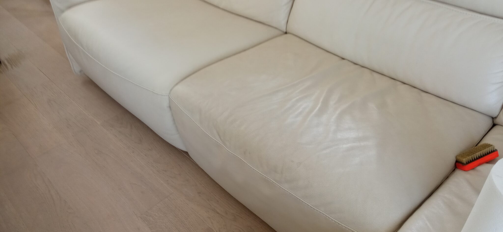 Fragment białej skórzanej kanapy z widocznym zabrudzeniem na siedzisku oraz szczotką do czyszczenia, leżącą obok rolki ręcznika papierowego na drewnianej podłodze.