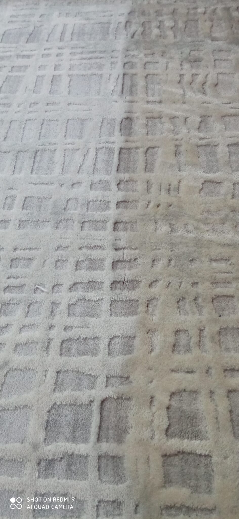 Fragment jasnoszarego dywanu z geometrycznym wzorem w kwadraty, widoczne różnice w odcieniach sugerujące obszar po czyszczeniu.