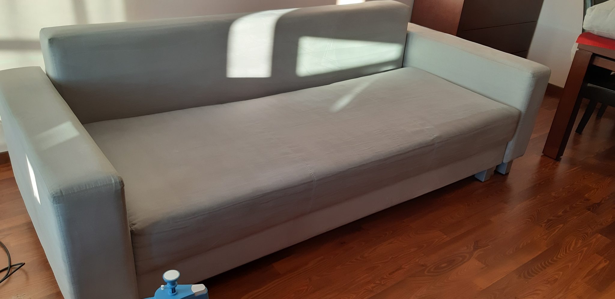 Jasnoszara sofa z widocznym urządzeniem do czyszczenia tapicerki na drewnianej podłodze, oświetlona naturalnym światłem słonecznym.