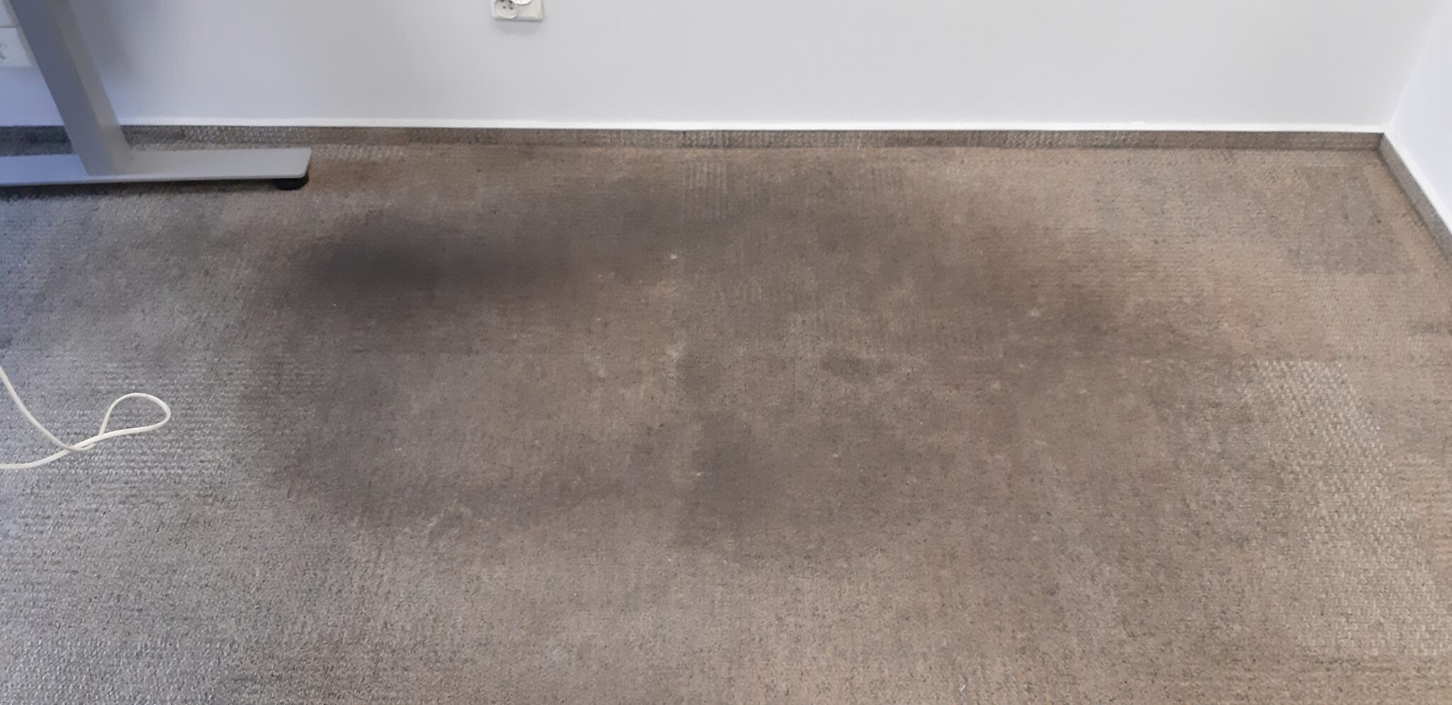 Szaro-beżowy dywan z widocznymi ciemnymi plamami zabrudzeń przy listwie przypodłogowej i nodze biurka, z kablem leżącym na powierzchni.