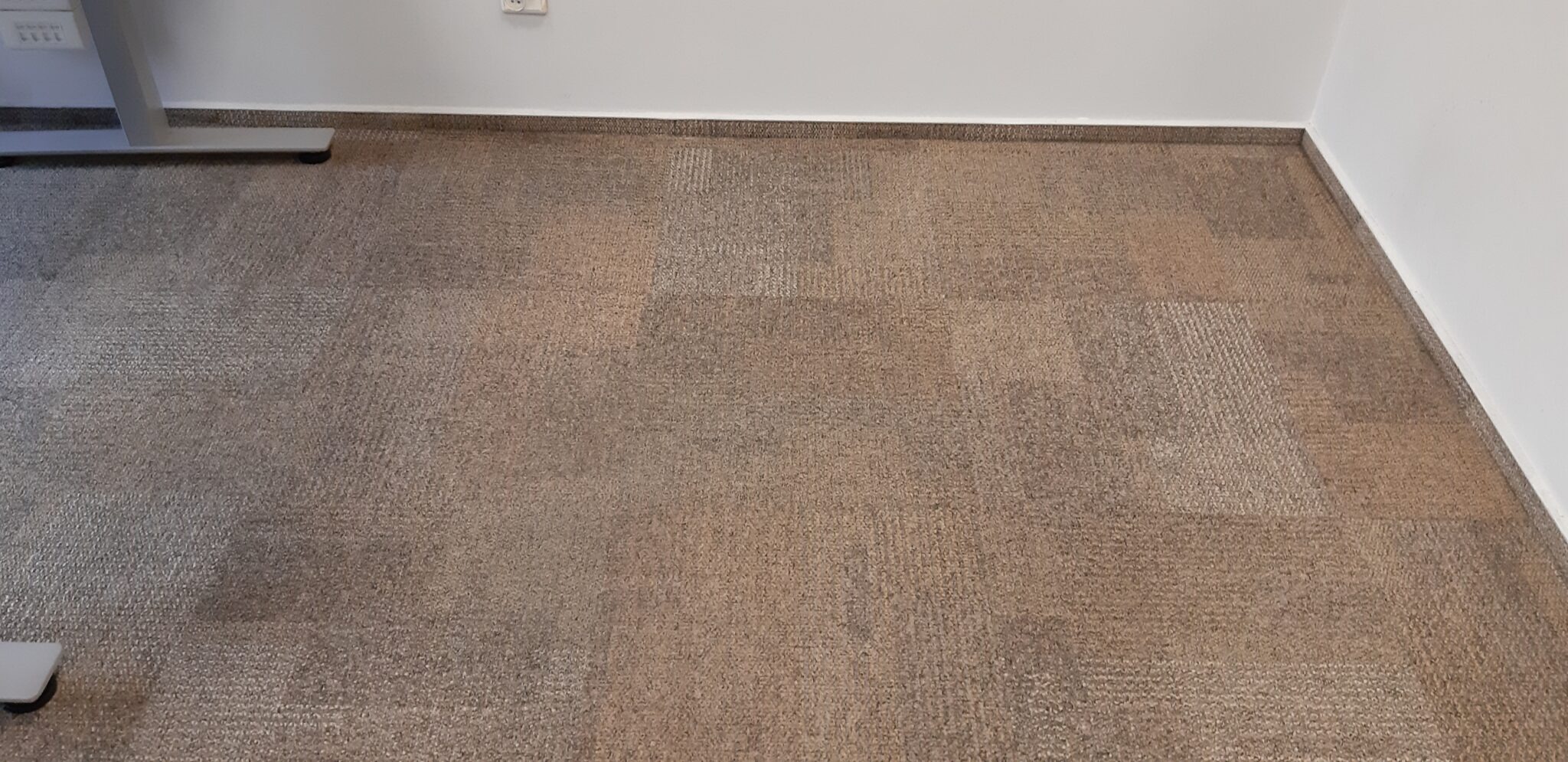Beżowa wykładzina dywanowa w biurze, widoczne ślady po czyszczeniu w postaci jaśniejszych kwadratów, fragment nogi biurka w lewym dolnym rogu.