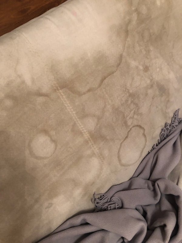 Beżowa tkanina z widocznymi, okrągłymi plamami i smugami, leżąca obok szarego koca z frędzlami.