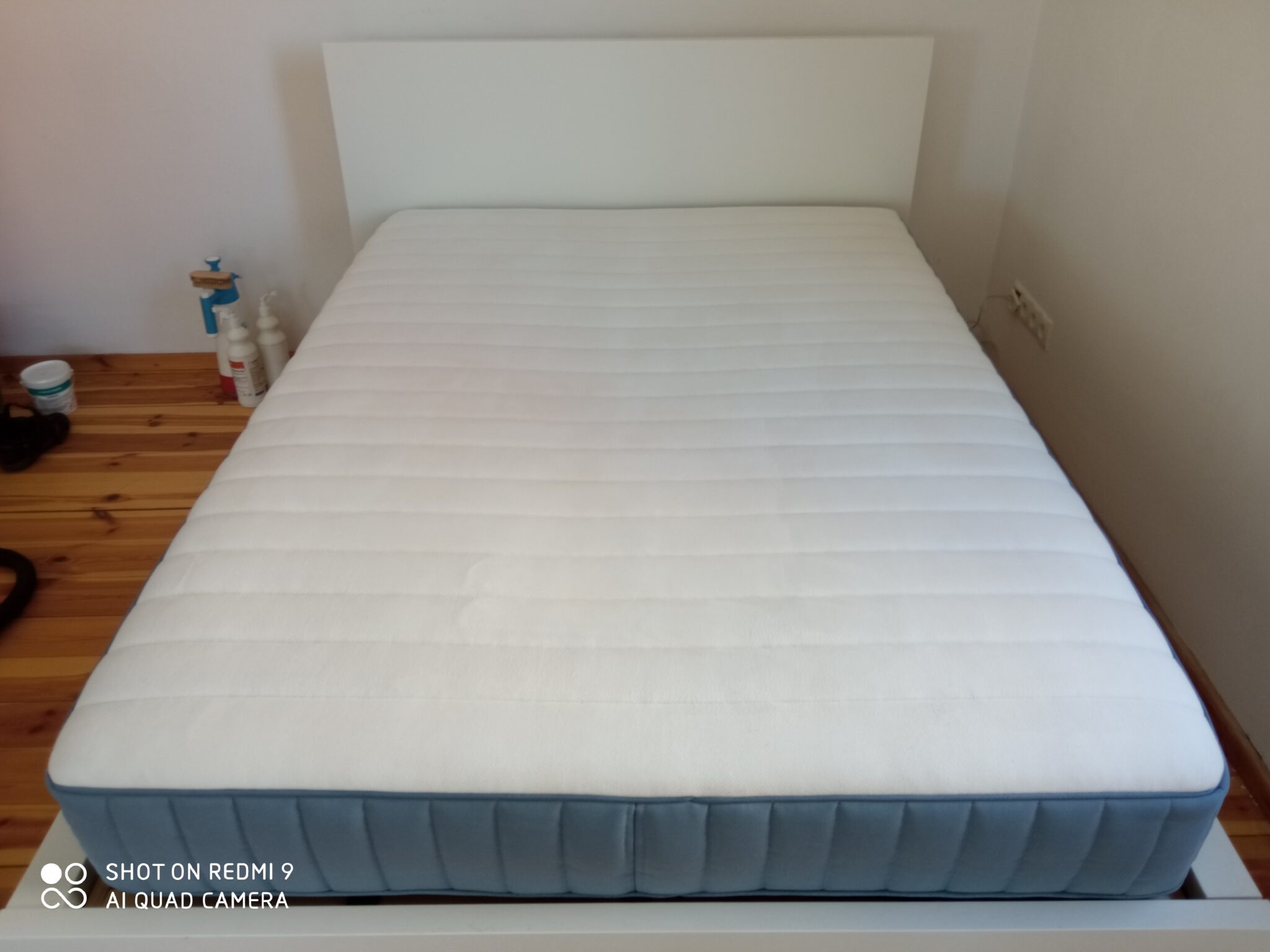 Nowy materac z białą powierzchnią i niebieskim obramowaniem leżący na białym łóżku, widoczne drewniane podłogi i ścianę w tle.