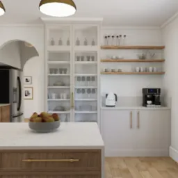 Jasna kuchnia z wyspą, na której leżą gruszki w szarej misce. W tle przeszklona szafka z zastawą, drewniane półki z naczyniami oraz ekspres do kawy i czajnik.