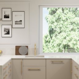 EMILIA MATYJASZCZYK ARCHITEKT WNĘTRZ - Jasna kuchnia z beżowymi szafkami, złotymi uchwytami i armaturą, okno z widokiem na zieleń, trzy czarno-białe zdjęcia w ramkach oraz ozdobna lampa nad blatem.