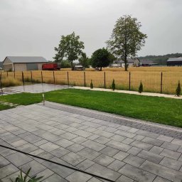 Electrostar kompleksowe usługi ogrodnicze - Widok na ogród z nowo ułożoną szarą kostką brukową, trawnikiem, młodymi tujami wzdłuż ścieżki, polem i zabudowaniami gospodarczymi w tle.