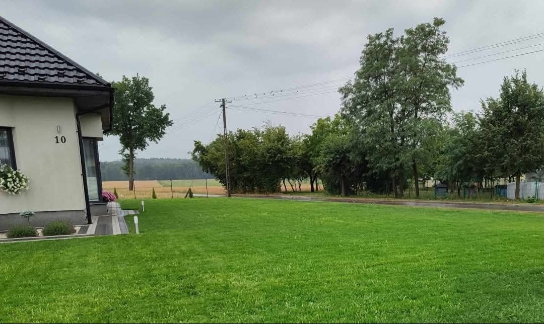 Starannie utrzymany trawnik przed domem jednorodzinnym z numerem 10, widok na pole w tle, pochmurne niebo.