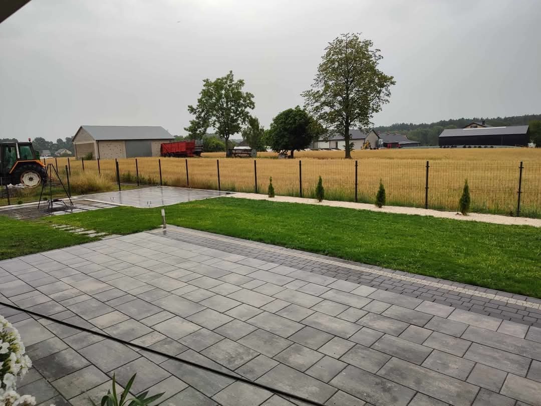 Widok na ogród z nowo ułożoną szarą kostką brukową, trawnikiem, młodymi tujami wzdłuż ścieżki, polem i zabudowaniami gospodarczymi w tle.