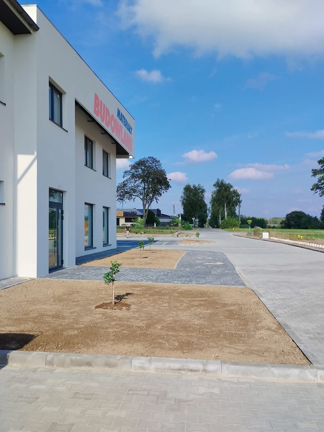 Nowo posadzone małe drzewka przed budynkiem z napisem 'MATERIAŁY BUDOWLANE', otoczone świeżo przygotowaną ziemią i kostką brukową.
