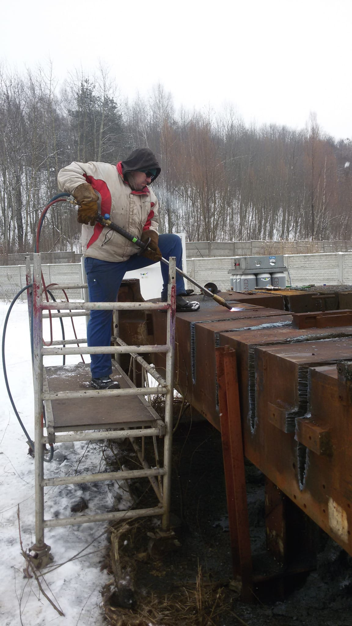 Pracownik w kurtce i rękawicach na metalowych schodach, używa palnika do cięcia grubej, zardzewiałej metalowej konstrukcji na zewnątrz w zimowy dzień.