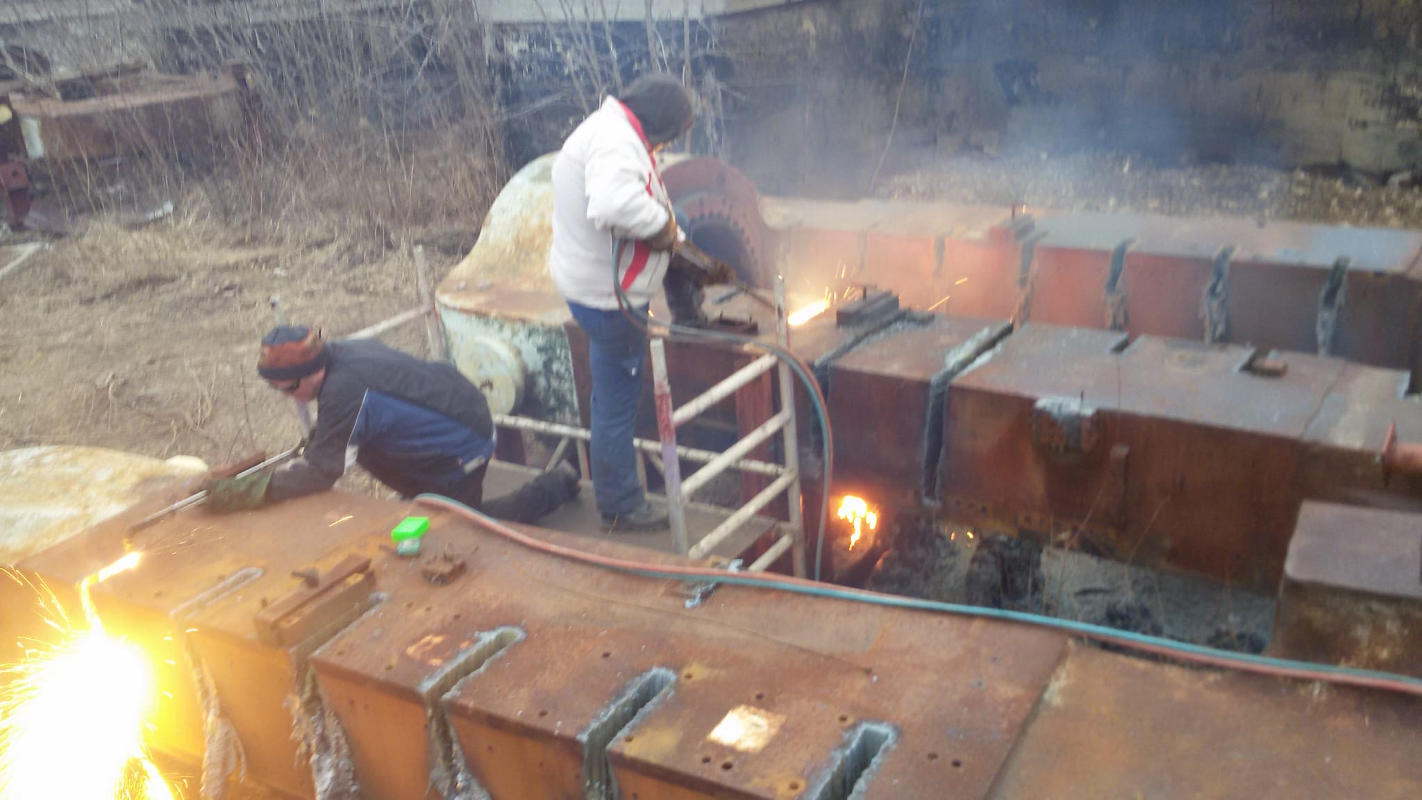 Dwóch robotników w roboczych ubraniach tnie palnikiem tlenowo-acetylenowym metalową konstrukcję na placu budowy, iskry i dym w powietrzu.