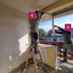 Netglow Amadeusz Bielecki - Dwóch mężczyzn w trakcie wyburzania ściany balkonowej za pomocą łomu, widok z wnętrza mieszkania, fragmenty ściany na podłodze, drabina oparta o ścianę.