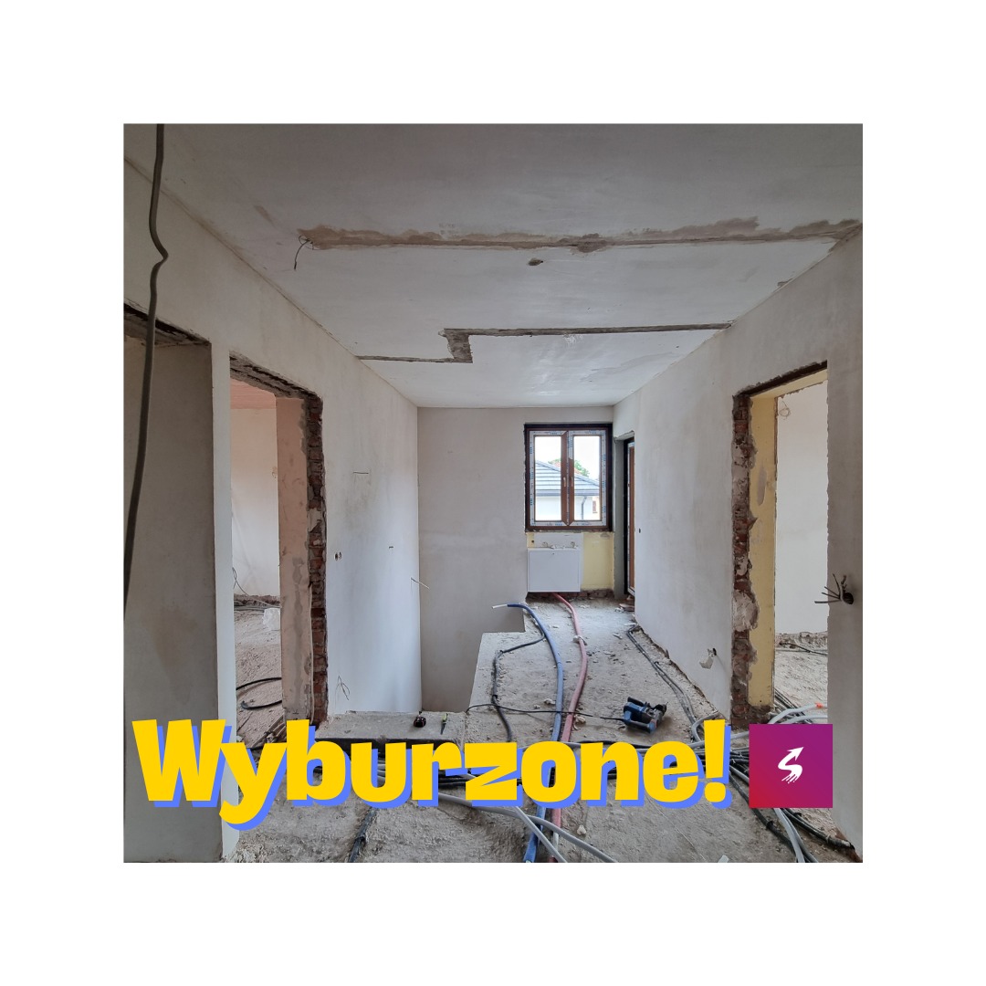 Wnętrze w trakcie prac, widoczne ślady po wyburzonych ścianach, odsłonięte cegły, wystające kable i rury, okno z widokiem na dach sąsiedniego budynku.