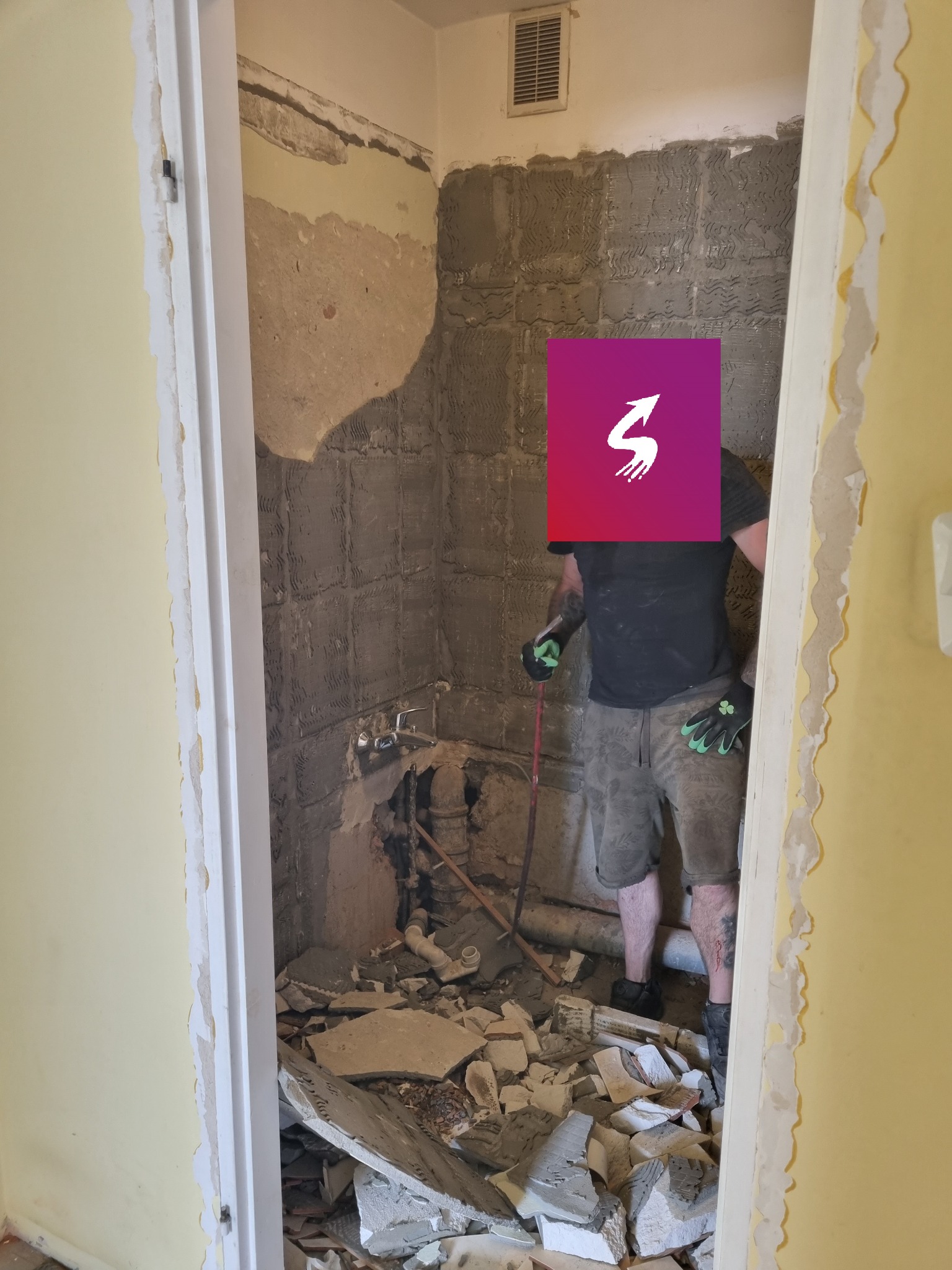 Wnętrze łazienki w trakcie wyburzania ścian, widoczne fragmenty płytek, rur i gruzu, pracownik w rękawicach ochronnych z narzędziem.