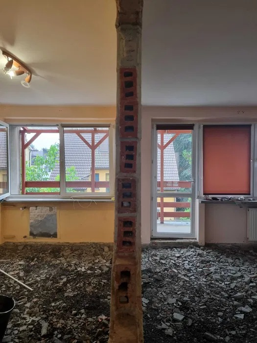Pozostałość po wyburzonej ścianie z cegły, widoczne gruzowisko na podłodze, okna z widokiem na zewnątrz, pomieszczenie w trakcie remontu.
