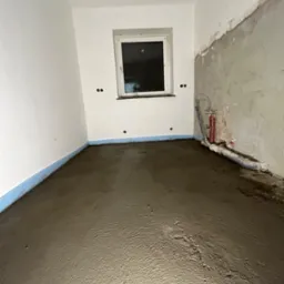 Pokój w trakcie wylewki samopoziomującej, widoczne przygotowane ściany, okno i wystające rury instalacji wodnej.