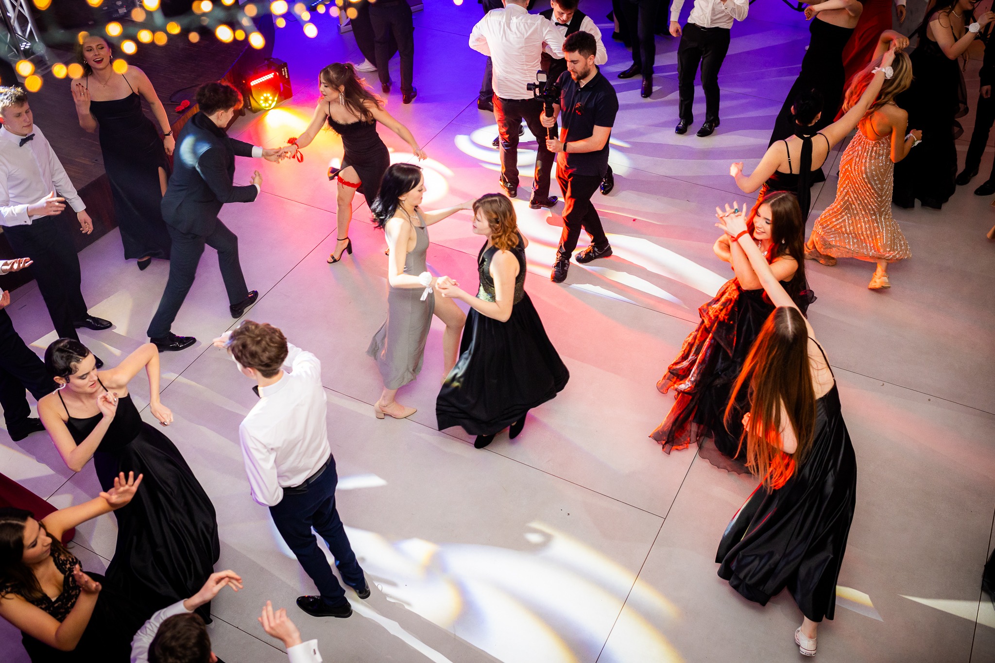 Widok z góry na bawiących się gości w eleganckich strojach na parkiecie oświetlonym kolorowymi reflektorami, fotograf uwieczniający momenty.