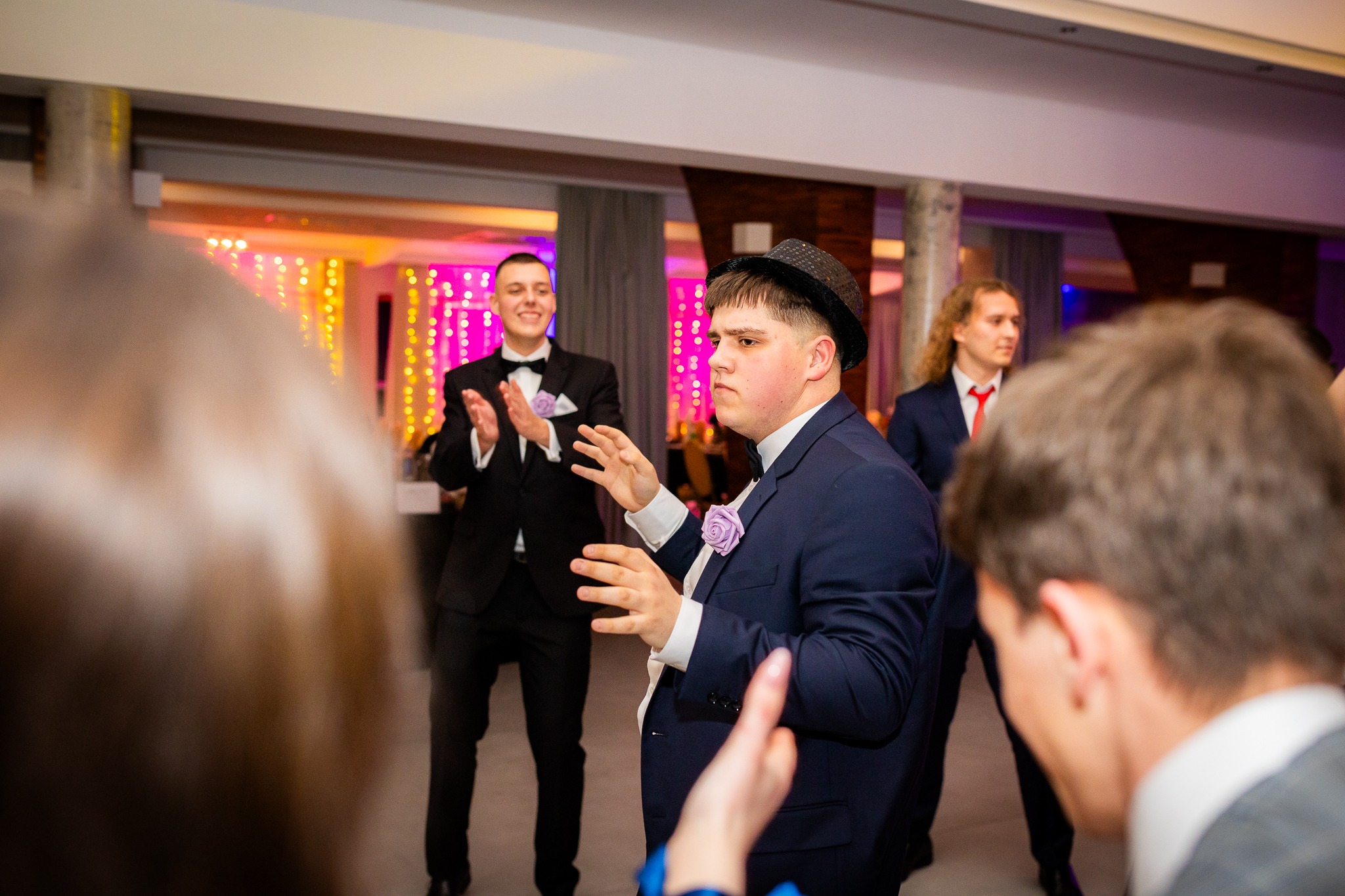 Mężczyzna w granatowym garniturze i czarnym kapeluszu tańczy na imprezie, w tle oświetlenie dekoracyjne i inne osoby w strojach galowych.