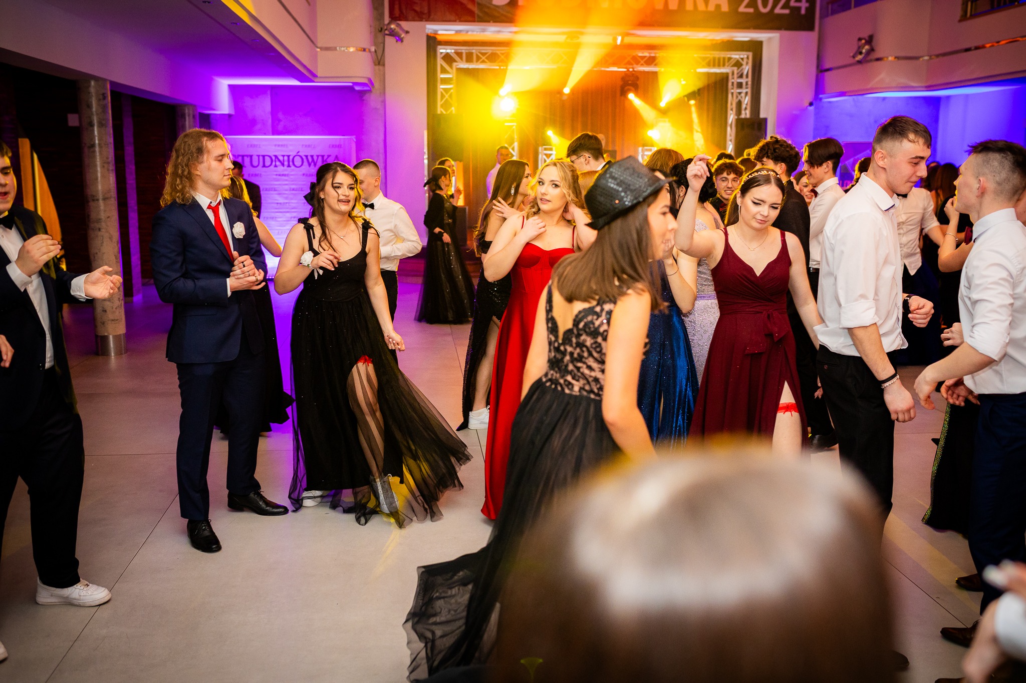 Grupa młodych ludzi w eleganckich strojach tańczy na studniówce, oświetlona kolorowymi światłami sceny. W tle widoczny napis 'Studniówka 2024'.