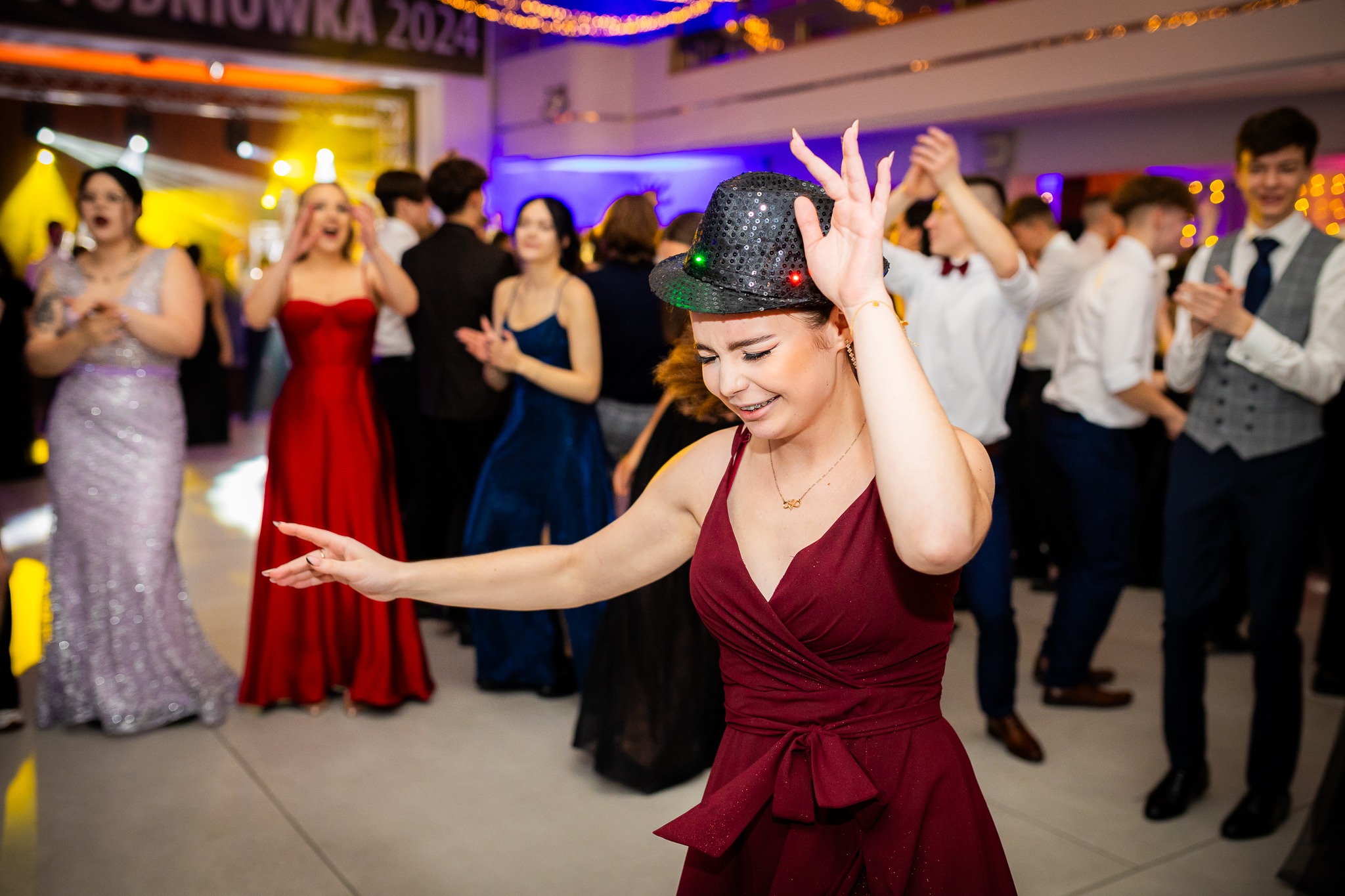 Uśmiechnięta dziewczyna w bordowej sukience i cekinowym kapeluszu tańczy na studniówce, w tle wiwatujący goście i dekoracje.