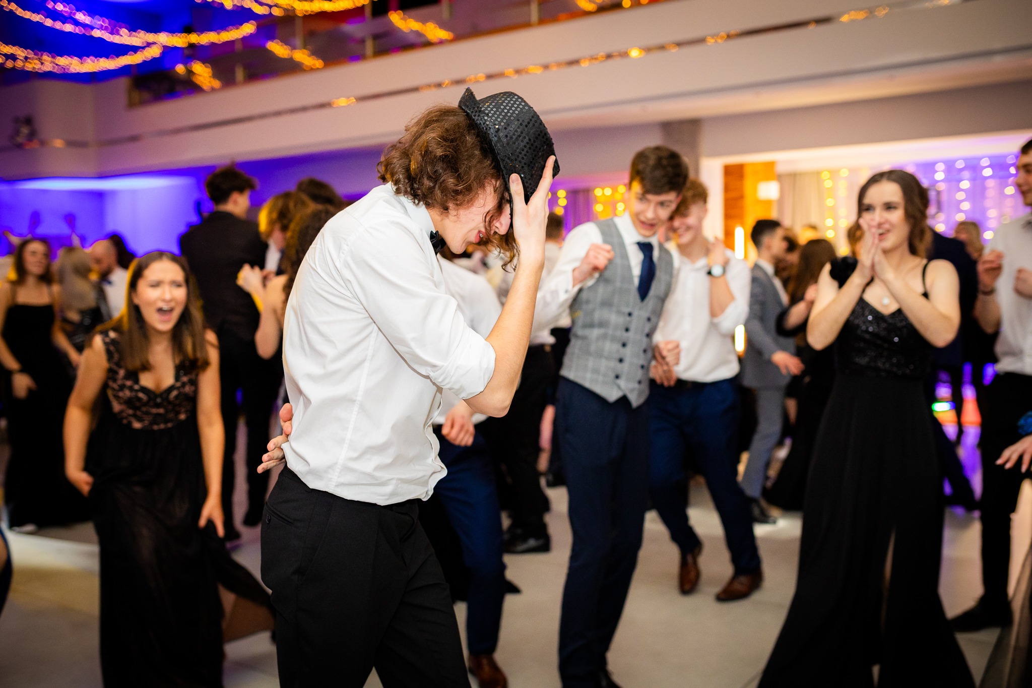 Rozbawiony mężczyzna w białej koszuli i czarnych spodniach unosi błyszczący kapelusz podczas tańca na parkiecie, w tle uśmiechnięci goście w eleganckich strojach i dekoracyjne oświetlenie.