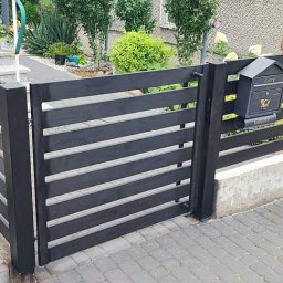 ParvisWelder - Nowoczesne, poziome, ciemnoszare ogrodzenie z furtką i skrzynką na listy, wykończone betonową podmurówką i kostką brukową, otoczone zielenią.