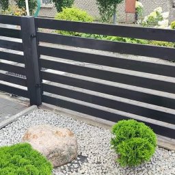 ParvisWelder - Nowoczesne ogrodzenie z poziomych, czarnych desek metalowych, otoczone białym żwirem, z widocznym fragmentem chodnika z kostki brukowej i dekoracyjnymi krzewami.