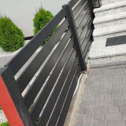 ParvisWelder - Nowoczesne, czarne ogrodzenie panelowe z czerwonym akcentem na słupku, przylegające do schodów wejściowych z lampą i kostki brukowej.