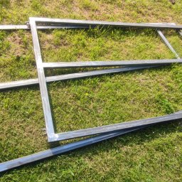 ParvisWelder - Surowe, spawane metalowe ramy na trawie, prawdopodobnie elementy bramy lub ogrodzenia, z widocznymi zawiasami i śladami po spawaniu.